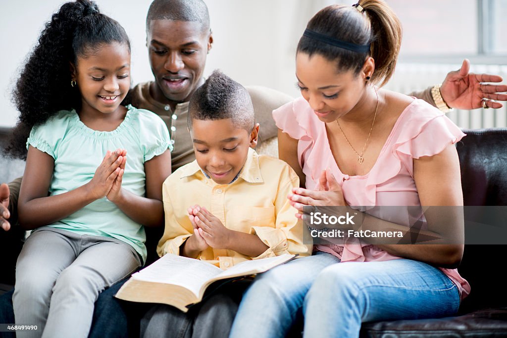Family Prayer An African-American family sitting on their sofa together, folding their hands for prayer with the bible open. Family Stock Photo