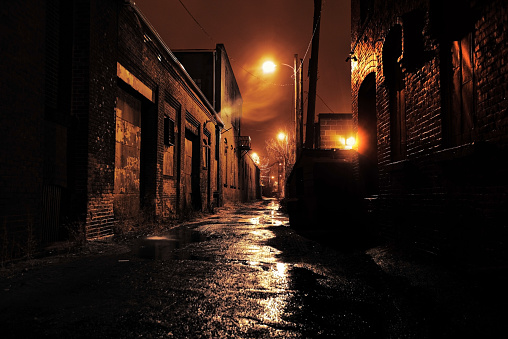 Long dark gritty alley between two old derelict buildings at night. Rain sodden pavement with eerie mist. Brickwork walls frame the ominous and dangerous inner city alleyway