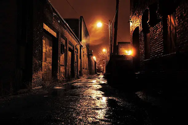 Long dark gritty alley between two old derelict buildings at night. Rain sodden pavement with eerie mist. Brickwork walls frame the ominous and dangerous inner city alleyway