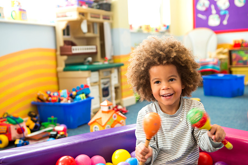 An image of a young boy happily playing in a ball pit, the boy is sitting down smiling surrounded by a colourful array of plastic balls. The boy holds a pair of maracas and looks like he is having a great time.