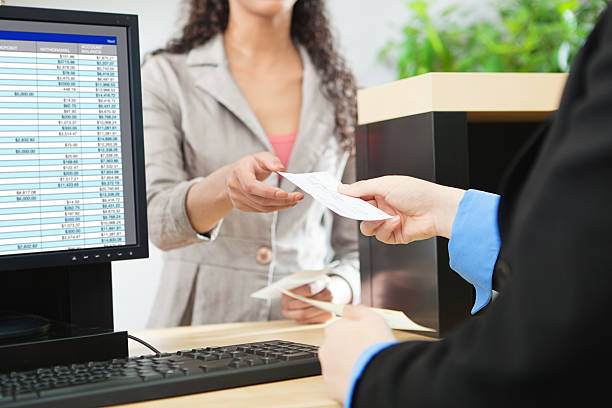 Bank Teller and Customer Banking Transaction in Retail Bank Counter Over-the-shoulder shot of a transaction between a bank teller and a customer in a retail bank. The teller is wearing a black suit and receiving a check from the tan-suited customer over the bank counter window. Photographed in horizontal format. bank teller stock pictures, royalty-free photos & images