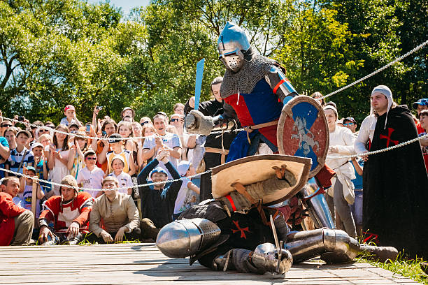 Tuttlingen, Baden-Wuerttemberg, Germany – 6.10.2022: Two knights in armor engage in a jousting duel on horseback at a medieval festival. One knight wields a lance and shield with a floral emblem, while the other raises a lance. Horses with caparisons are part of the scene, with a wooden barrier separating them. The event takes place before an audience, including families, under tents and banners, suggesting a festive and historical atmosphere. Lush greenery in the background adds to the setting's medieval ambiance.