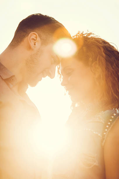 Epic close up portrait of a adorable european female with sunglases black eyes loolking into camera smiling while is kissded by her man against beautiful sunset with sunflares .