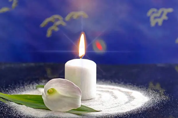 a single white burning candle and stands in white sand. Next to it is a white calla flower. The background is blue