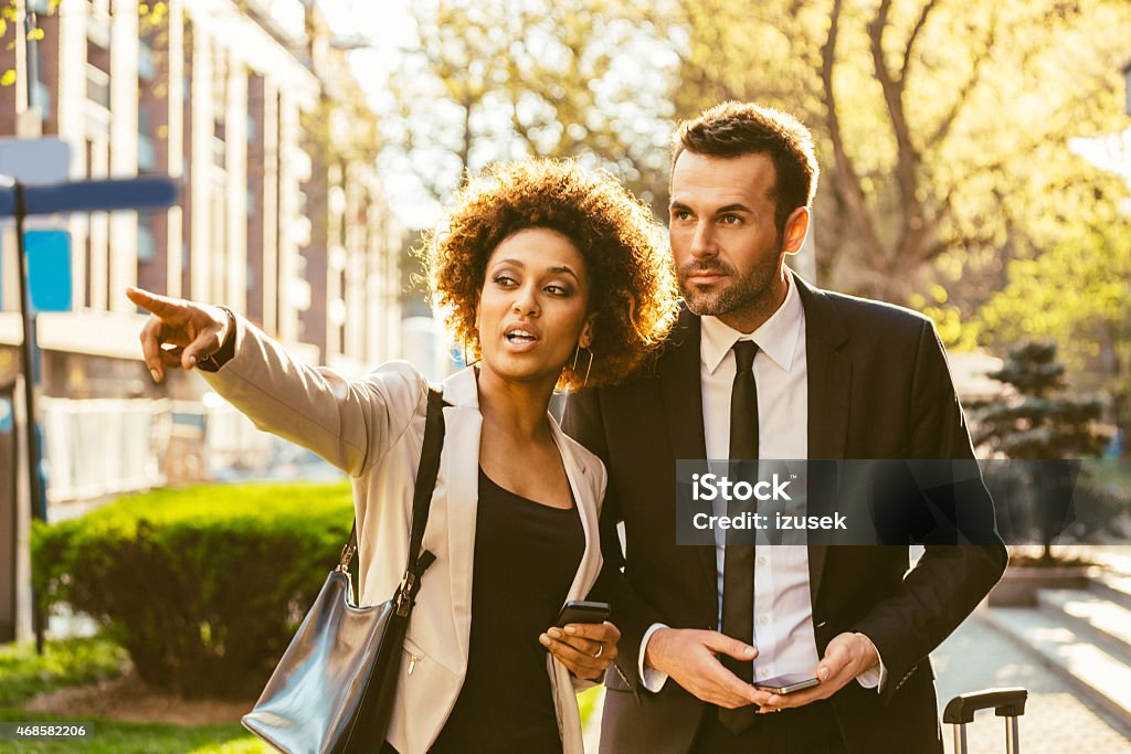 Outdoor portrait of two business people with smart phones Portrait of businessman and afro american businesswoman in elegant outfits holding smart phones in hands, standing outdoor at sunset. Woman pointing with index finger a direction. Real Estate Agent Stock Photo