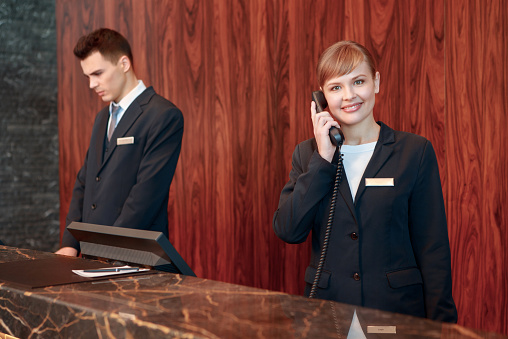 Reception on the phone. Young smiling receptionist in black uniform answering the call at the hotel counter