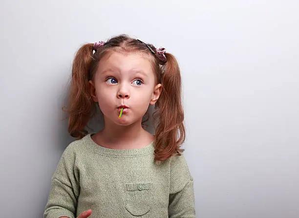 Funny thinking kid girl eating lollipop and looking up on grey background