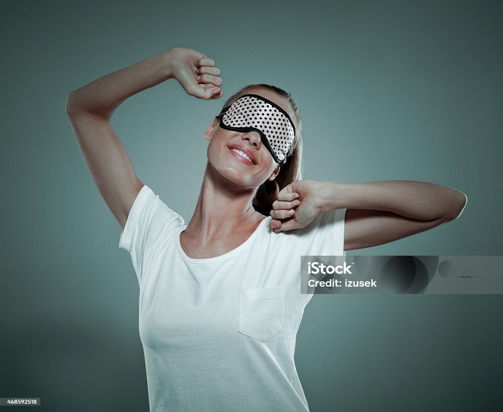 Smiling young woman wearing sleeping mask on her eyes Conceptual portrait of smiling blonde young woman wearing white t-shirt and sleeping mask on her eyes. Standing against grey background and stretching her amrs. Studio shot. Eye Mask Stock Photo