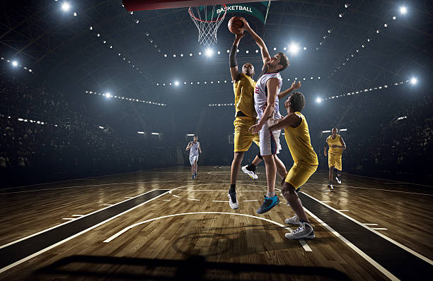 Basketball game Low angle view of a professional basketball game. A player is in mid air holding ball about to score a slam dunk, but the player from the opposite team is ready to block him.  A  game is in a indoor floodlit basketball arena. All players are wearing generic unbranded basketball uniform. offense sporting position photos stock pictures, royalty-free photos & images