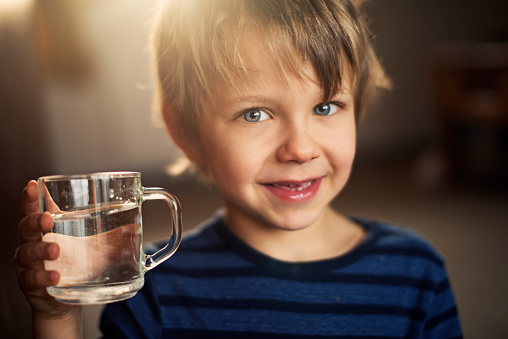 Closeup portrait of a little boy holding a glass of water. The boy is wearning blue blouse and smiling to the camera. The boy is aged 5 and is backlit by the morning sun from the window behind him.