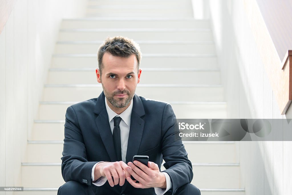 Businessman using a smart phone Portrait of confident businessman wearing dark suit sitting on white stairs in an office and using a smart phone, looking at camera. 2015 Stock Photo