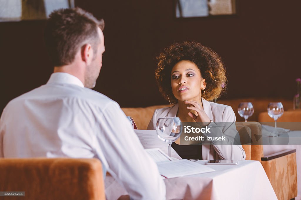 Two business people at lunch in the restaurant An afro american businesswoman and caucasian businessman having lunch or dinner in restaurant, sitting at the table, reading manu and talking about ordering. Business Lunch Stock Photo