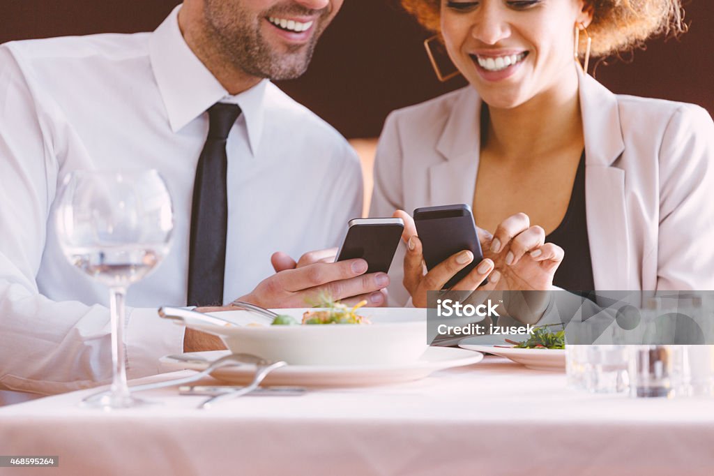 Businessman and businesswoman using smart phones at lunch Afro american businesswoman and caucasian businessman having lunch or dinner in the restaurant, using smart phones together. Close up of smiling faces and hands. Wineglass and plates on the table. Bar - Drink Establishment Stock Photo