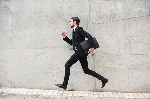 Happy young man in formalwear holding briefcase while running in front of the concrete wall
