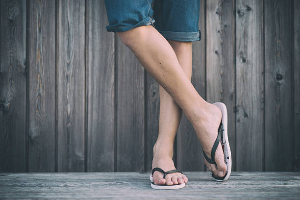 Man's Summer Legs with Flip Flops Thigh-down shot of a young man wearing denim shorts and flip flops, chilling out in summer on the boardwalk. The legs are crossed. thong stock pictures, royalty-free photos & images