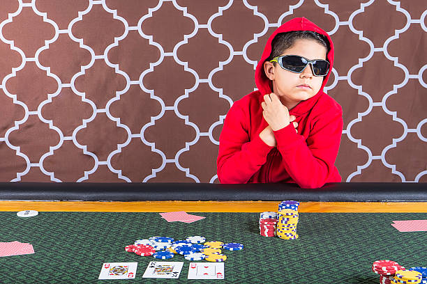 Young boy playing poker at a table A young boy sitting at a poker table with a hoodie and sunglasses gambling playing cards with a brown background child gambling chip gambling poker stock pictures, royalty-free photos & images