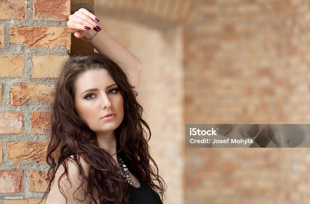 Portrait of beautiful woman in black Attractive young woman leaning on brick pillar. She has black T-shirt, her left hand over head, long brown hair and looking dreamily into camera. Shooting with Canon EOS 5D Mark II, selective focus. 20-24 Years Stock Photo