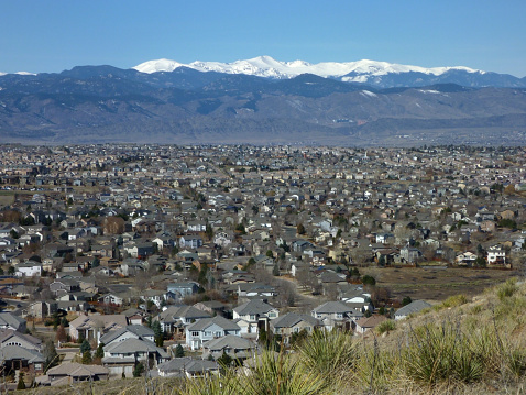 Thousands of homes and condos along the Front Range community of Highlands Ranch stand packed tightly together in front of the snow covered Mount Evans in the Rocky Mountains of Colorado with yuccas and grasslands in the foreground.
