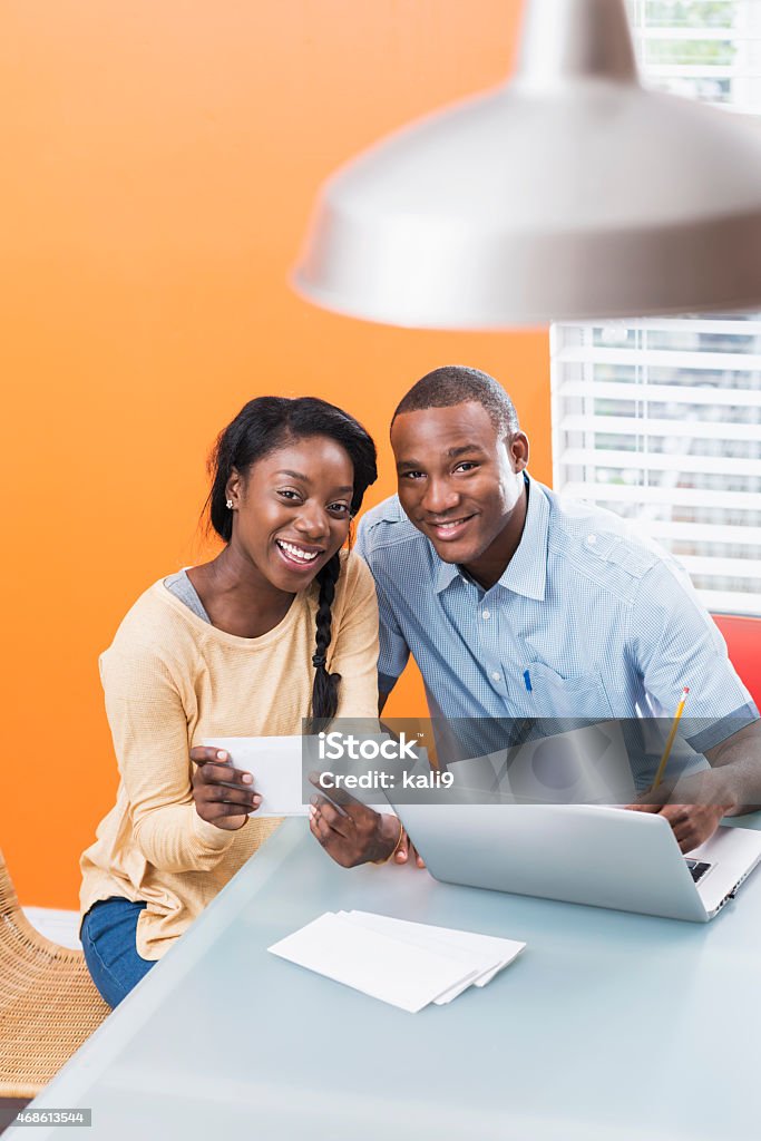 Young black couple paying bills at home High angle view of a young African American couple at home, paying bills online using a laptop computer.  They are sitting at a table in an orange room, smiling at the camera. 20-29 Years Stock Photo