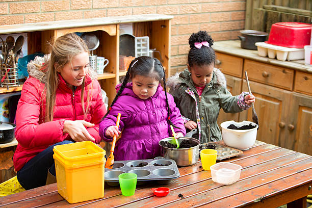Messy Mud Pies A horizontal image of two children outdoors using a big mental pan to make mud pies, while a young woman supervises. The children are both wrapped up warm in coats and are contently playing nursery stock pictures, royalty-free photos & images