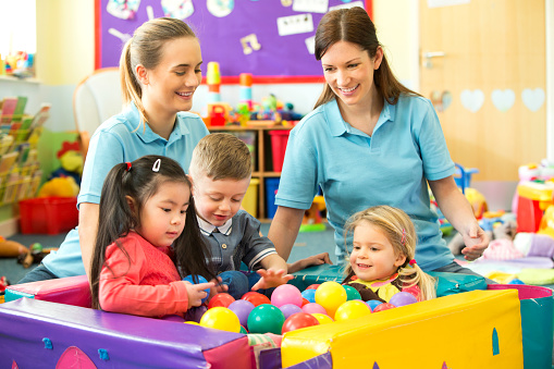 A candid image of three children in a ball pool playing and laughing, behind them sit two nursery teachers in blue polo shirts supervising and smiling. The nursery is a colourful scene of toys and mess.