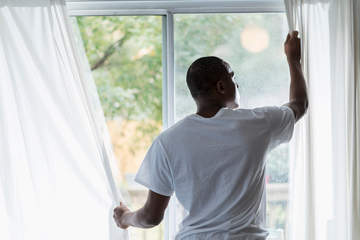 Rear view of a young black man opening the white curtains to let in the morning sunlight.  His face is visible enough to see his serious expression.