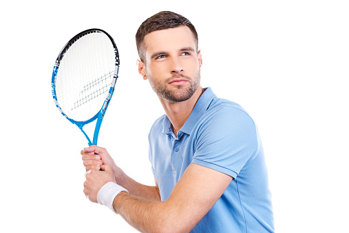Confident young man holding tennis racket and looking concentrated while standing against white background