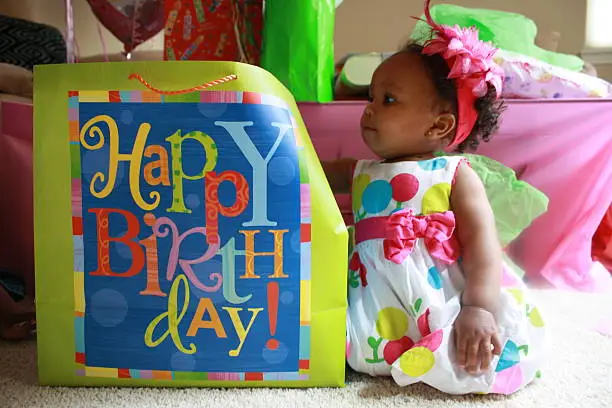 1 year old multi-racial birthday girl looking at large birthday gift bag.