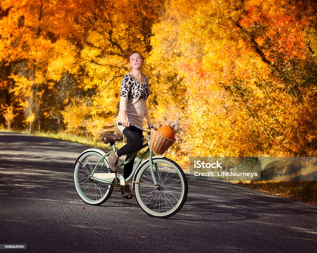 Young Woman Riding Her Bike On A Paved Autumn Pathway Young Woman Riding Her Bike On A Paved Autumn Pathway .  Her basket is full of punpkins and corn.  She is delightfully oblivious to the photographer, enjoying her hair blowing in the wind as she rides. 2015 Stock Photo