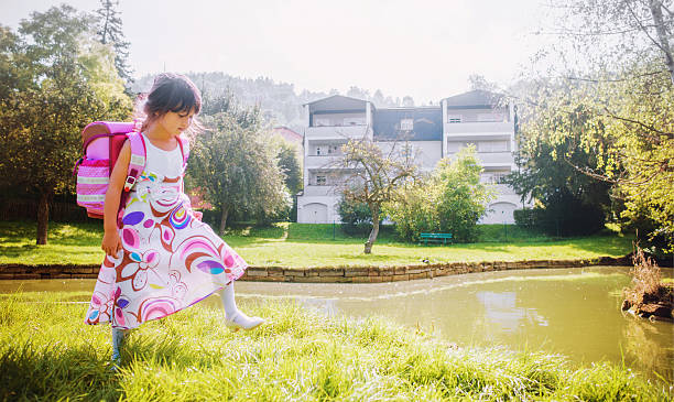 6 year old German-Japanese school girl wearing proudly a beautiful summer dress and her satchel, daydreaming and strolling back home through a park after her first day at elementary school.