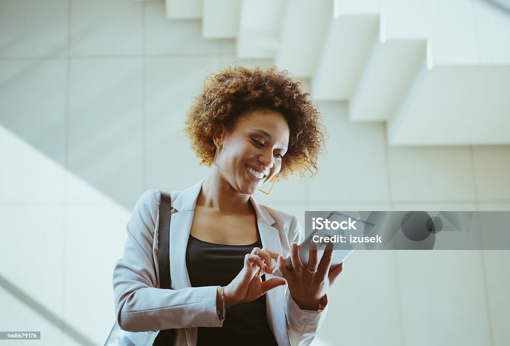 Afro american woman using a digital tablet Happy afro american businesswoman wearing jacket standing against white wall and stairs in an office and using a digital tablet. Ecstatic Stock Photo