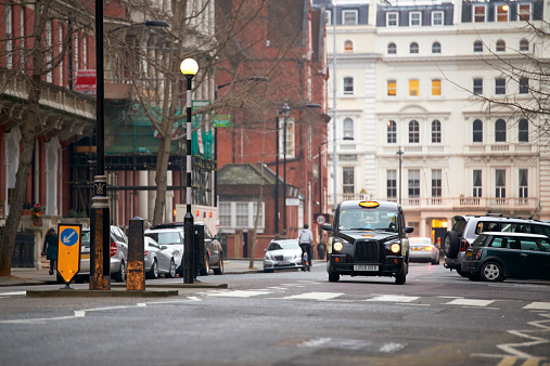 Famous London black cab in residential road in early evening. There are cars parked on the sides, and traditionally British residential buildings in the background.