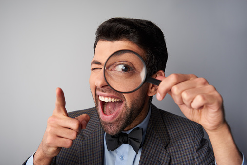 Funny picture of positive casual young man wearing jacket and bow tie. Man smiling, holding magnifier near eye and pointing at camera