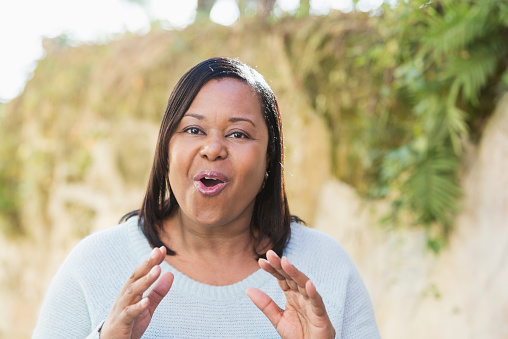 Face of a confident, mature, African American woman standing outdoors, talking to the camera.  Her mouth is open, smiling and speaking at the same time, and she is gesturing with her hands.