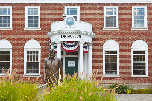 Hyannis, MA, USA - September 16, 2014: JFK (John Fitzgerald Kennedy) Museum in Hyannis, Cape cod, Massachusetts, USA. Tall green grass and bronze Statue of JFK are in foreground. The image gives an impression that the JFK is walking through the tall grass. Red brick museum buiding is in background. Museum portico shows the inscription 