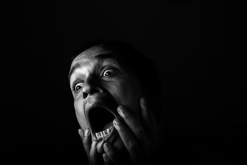 A black and White portrait of a man in extreme despair and sorrow. The man has his eyes wide open and eye-balls protruding out. He is screaming in despair and so his mouth is also wide open. Very dramatic lighting and expression in this image. The background is jet black.