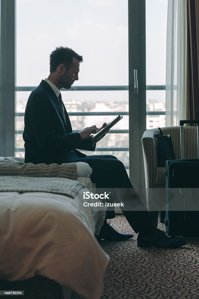 Businessman sitting in hotel room and using digital tablet Silhouette of businessman wearing suit sitting on bed in hotel room and using a digital tablet with city scape from the window. 2015 Stock Photo