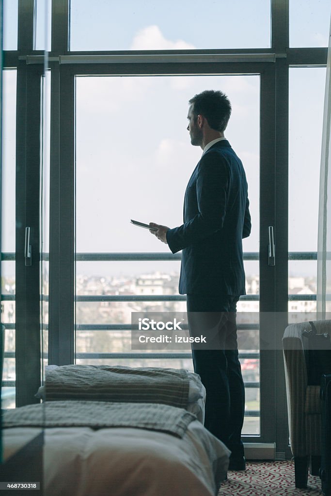 Businessman standing in hotel room and holding digital tablet Businessman wearing suit standing in hotel room, holding a digital tablet and looking throught out the window at the cityscape. 2015 Stock Photo
