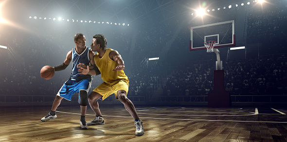 Low angle view of a professional basketball game. A player is trying to block  the opposite team player with a ball. A game is in a indoor floodlit basketball arena. All players are wearing generic unbranded basketball uniform.