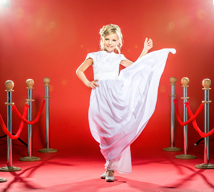 Beautiful little girl in a white eveningwear is walking on a red carpet holding a hem of her dress in the spotlights