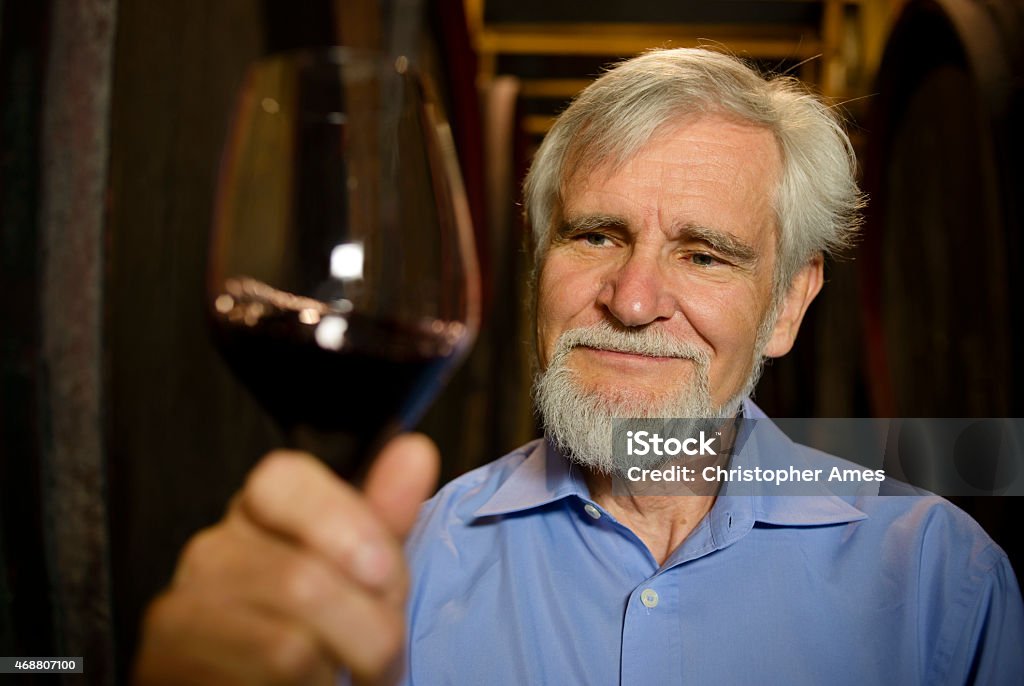 Senior Male Winemaker With Glass of Red Wine in Cellar A senior man with grey hair and beard gives a satisfied look at a glass of red wine he holds aloft. He is standing in a wine cellar, in front of two rows of wooden wine barrels. The man appears to be an experienced wine maker and probably owner of the vineyard or winery. Camera: 36MP Nikon D800E. 2015 Stock Photo