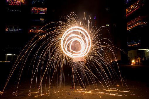 A man is playing with a firecracker on the occasion of Diwali in India. The man is invisible and hidden behind the firecracker. In the background, multi-storied residential buildings are seen which have been illuminated with lights to mark the occasion of Diwali. Diwali is the most auspicious religious festival of Hindus.