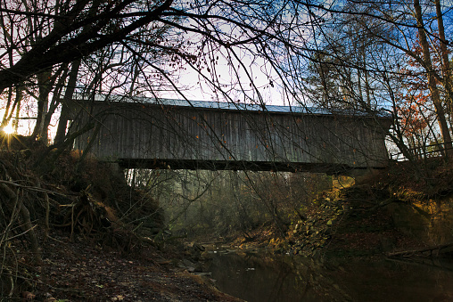 Wooden truss covered bridge, overlooking a creek in the woods, on a warm autumn morning.