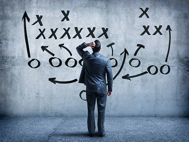 Businessman looking up at strategy diagram on wall A rear view of a businessman looking up at an American football diagram written on a large concrete wall. He is standing with his hand on his head as he tries to memorize all the different facets of the strategy. A blue color scheme dominates the image. american football game plan stock pictures, royalty-free photos & images