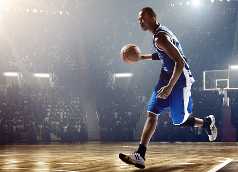 Low angle view of a professional basketball game. A player is running with a ball. A game is in a indoor floodlit basketball arena. All players are wearing generic unbranded basketball uniform.