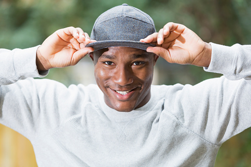Portrait of a handsome young African American man outdoors.  He is staring at the camera with a big smile on his face, holding the bill of the cap on his head.  He is happy and confident.