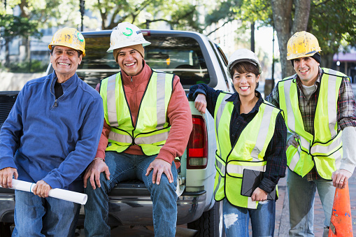 A group of four diverse workers wearing hard hats and yellow safety vests with pickup truck on city street.  They are a crew of construction or utility workers, smiling at the camera.  One of the workers is a woman and two are Hispanic.