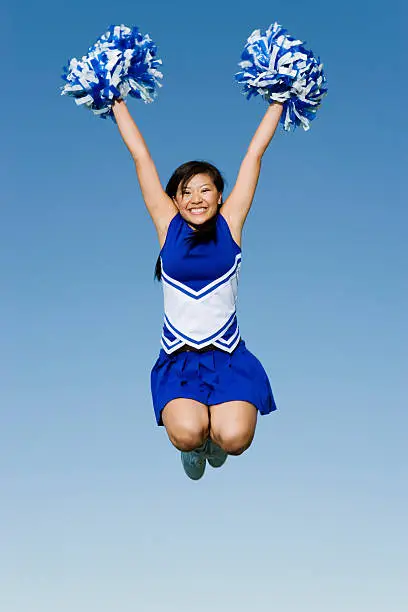 Full length of excited cheerleader with pompoms in midair against blue sky