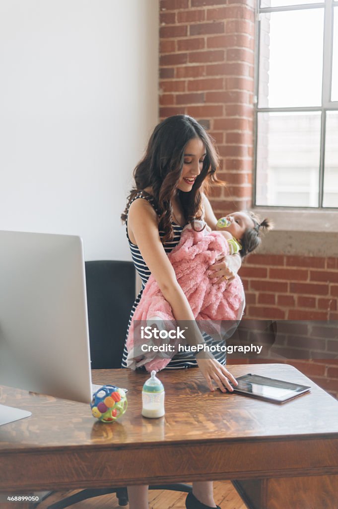 Working Mother Mother holding child while working on tablet at desk. 2015 Stock Photo