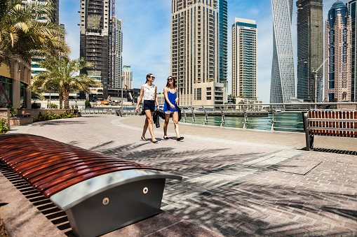 Two european friends meeting and greetings in the streets of central Dubai Marina during a sunny day in the summer. They are drinking coffee using a disposable cup and walking on the promenade.